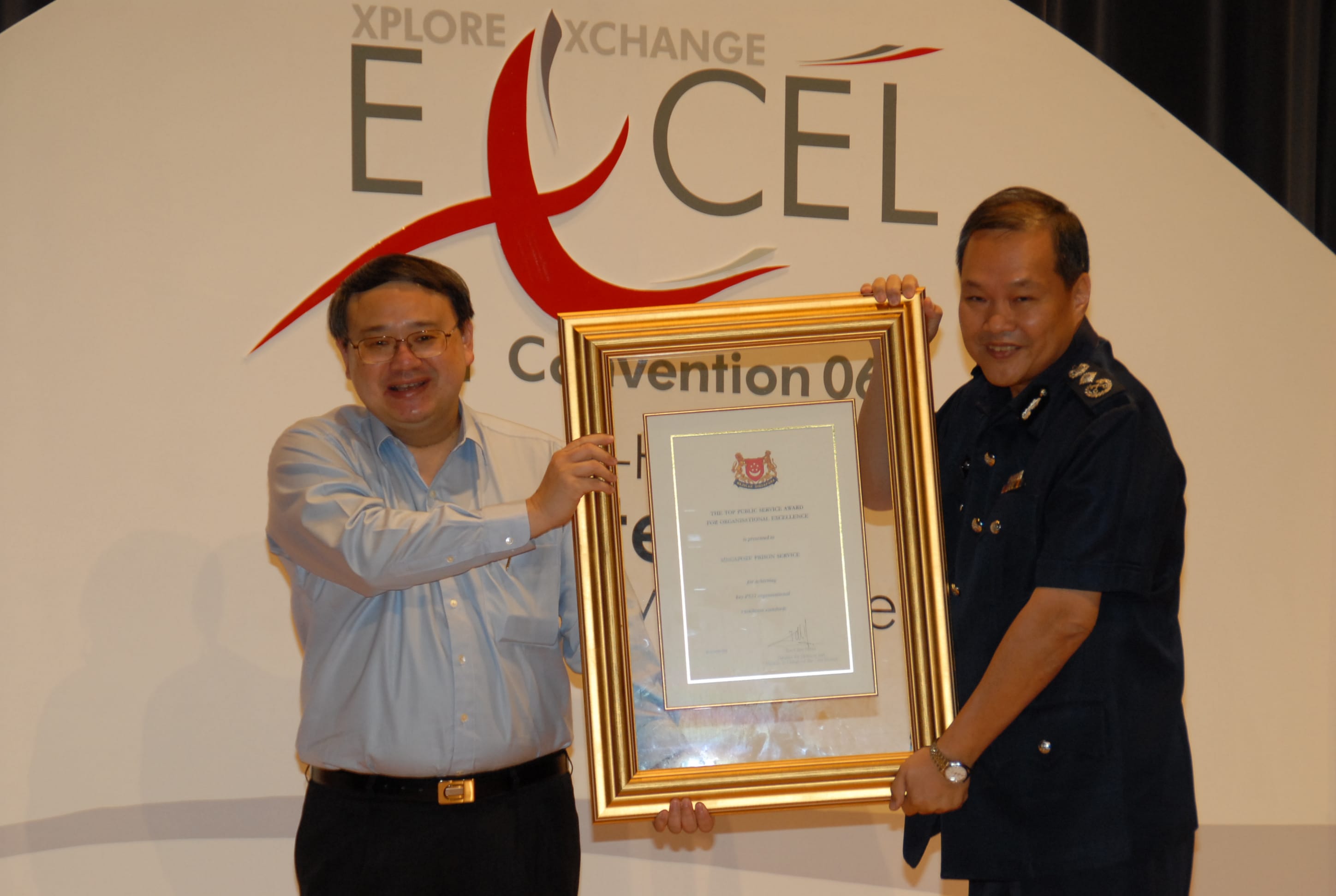 Two individuals holding a large framed certificate in front of a sign that reads "EXCEL Convention 06".
